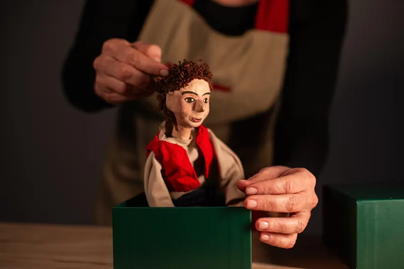 Mãos ajeitam o cabelo de lã do boneco enquanto ele emerge da caixa verde apoiada em uma mesa de madeira.
