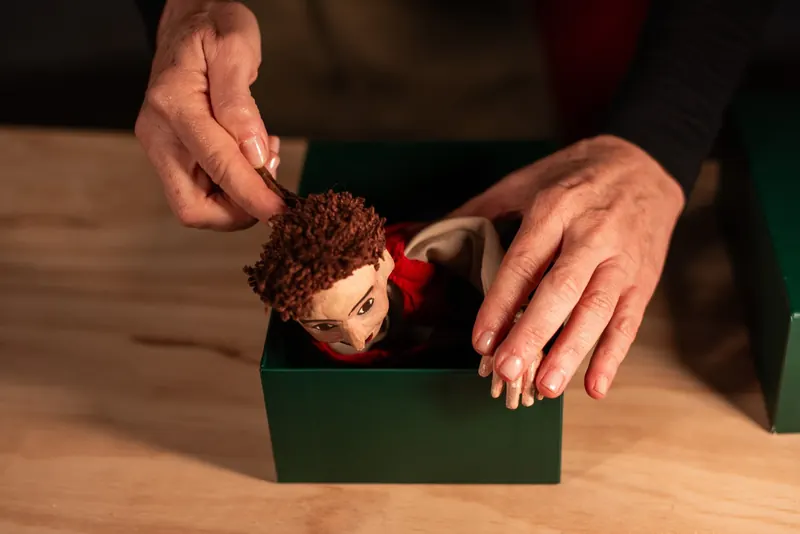 Mãos acomodam o boneco de cabelo castanho cacheado de cabeça para baixo dentro de uma caixa verde apoiada em mesa de madeira.