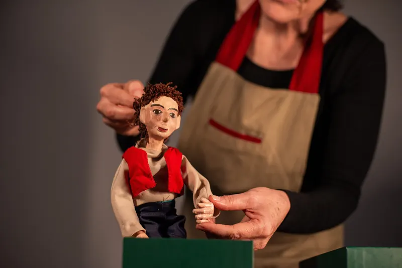 Boneco de colete vermelho e calça jeans é posto em pé sobre a caixa verde, sustentado pelas mãos da manipuladora.