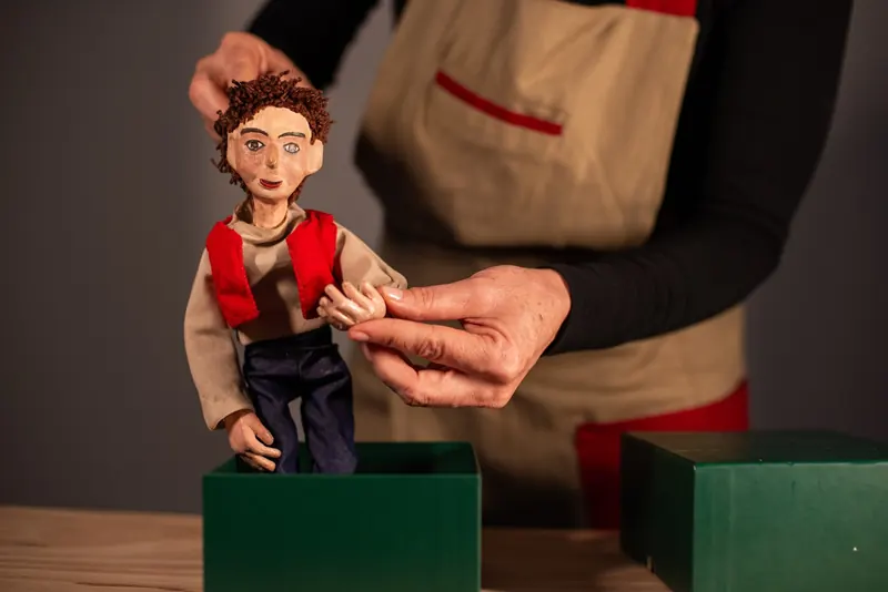 Manipuladora apoia o boneco em pé sobre a caixa verde e ajusta o braço dele, preparando o personagem para a cena.