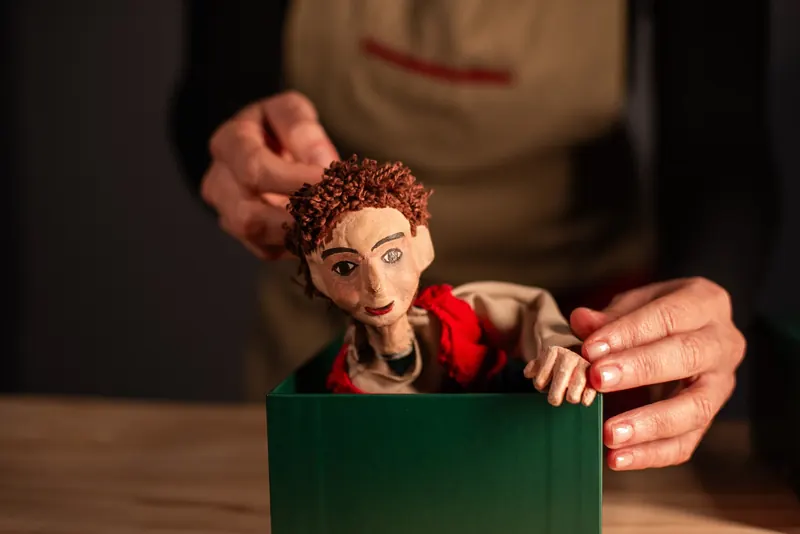 Mãos retiram com cuidado de uma caixa verde um boneco de cabelo castanho cacheado e colete vermelho.