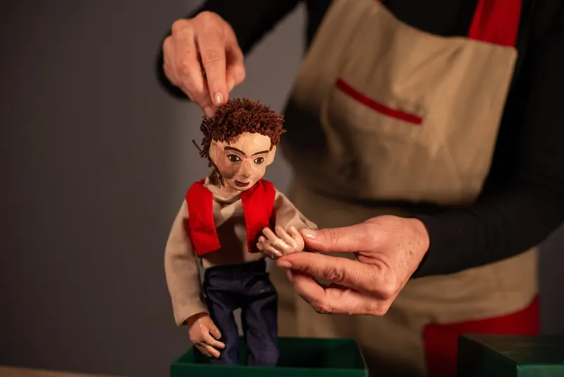 O boneco aparece em pé sobre a caixa verde, de corpo inteiro, com as mãos da manipuladora apoiando o braço.