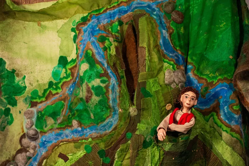 Detalhe do cenário de feltro visto de perto: um rio azul serpenteia entre manchas de verde, pedras de lã e retalhos costurados.