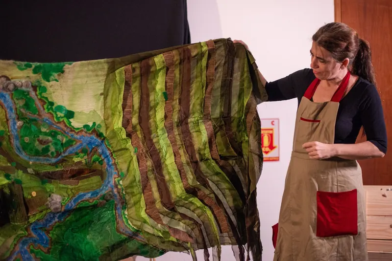 Manipuladora de avental sustenta com o braço erguido um amplo painel de feltro tingido em verdes e marrons, que representa a paisagem da peça.