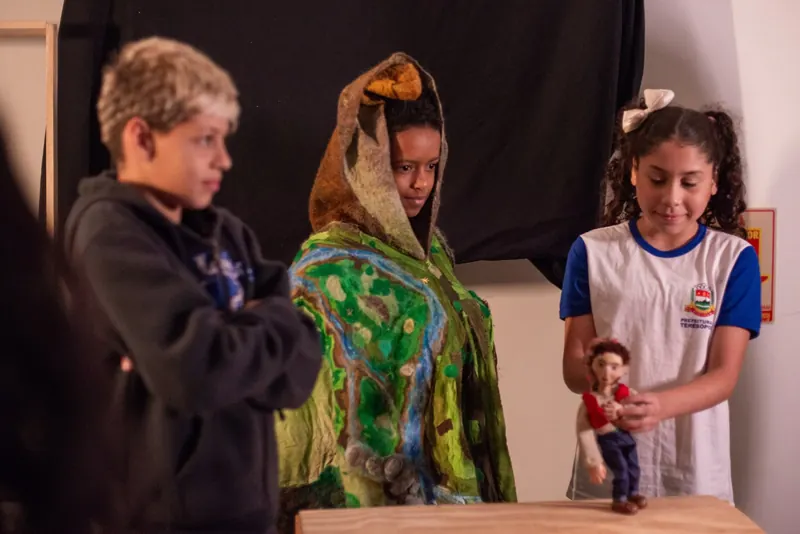 Estudantes experimentam elementos do espetáculo: um veste a capa de feltro do cenário e outra segura um dos bonecos.