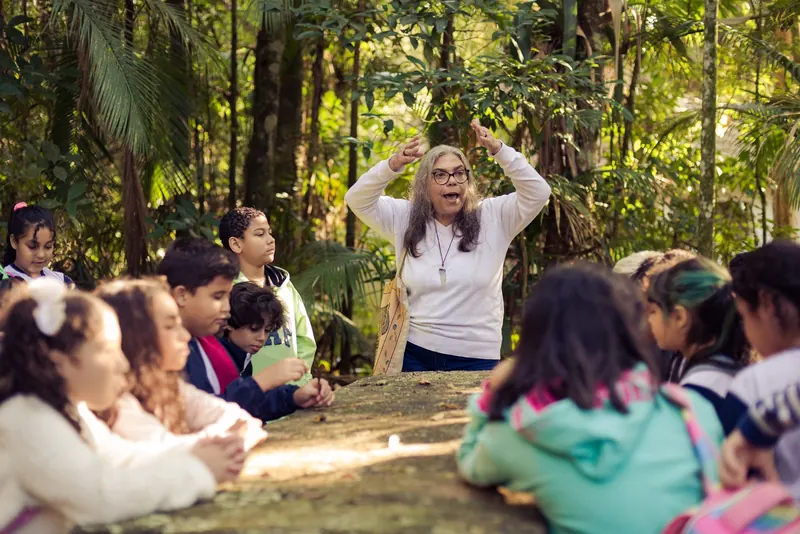Educadora ergue as duas mãos ao lado da cabeça, imitando um animal, diante de estudantes sentados em roda na mata.