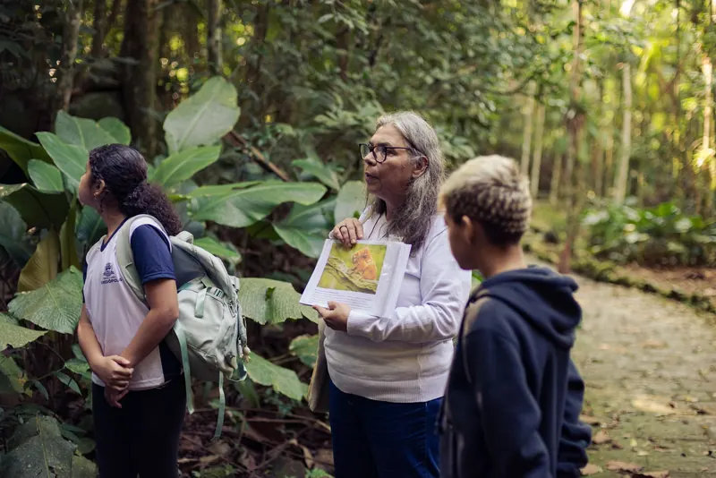 Educadora conversa com dois estudantes durante a caminhada, segurando o material impresso de apoio.