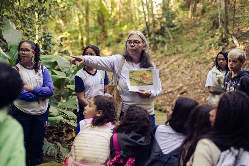 Educadora aponta para a copa das árvores enquanto segura uma prancha com a foto de um animal, cercada por estudantes.