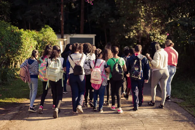 Turma de estudantes caminha de costas por um caminho pavimentado dentro da unidade de conservação, entre árvores e arbustos, no fim da tarde.