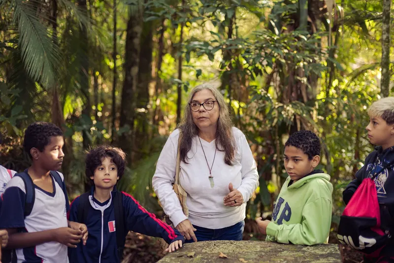 Educadora conversa com estudantes durante uma parada da trilha, cercada por vegetação densa.