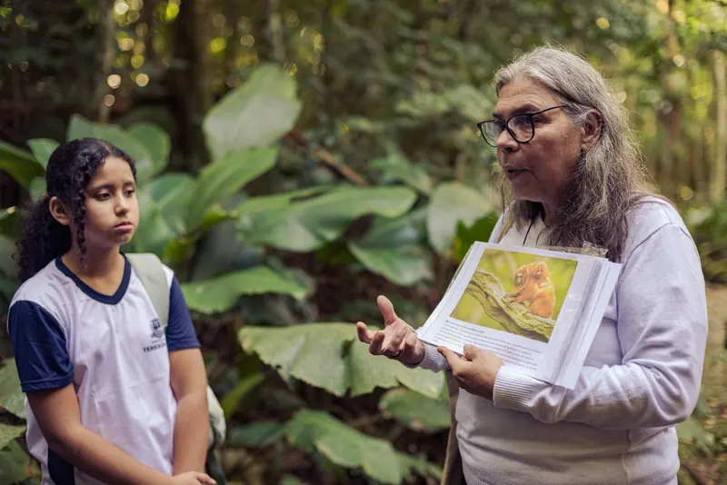 Educadora apresenta a uma estudante uma prancha com a foto de um mico-leão-dourado, em meio à vegetação.