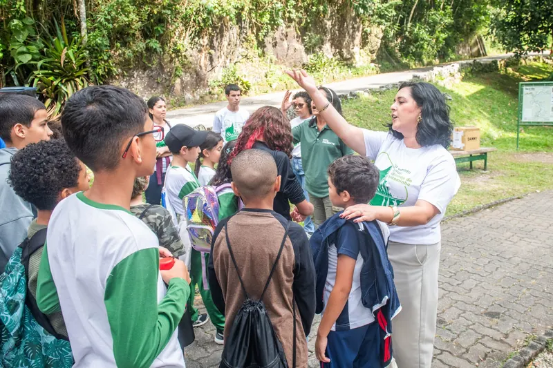 Educadora de camiseta do projeto conversa e gesticula para um grupo de estudantes reunido na área de recepção do parque.